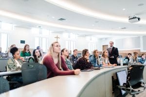 students facing the front of a bright and sunny classroom, looking at some kind of a classroom materials, while man in suit to the right speaks to them