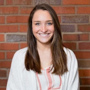 woman with brown hair and blue eyes looking at the camera, wearing a white shirt in front of a brick wall 