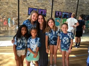 woman with brown hair posing with 5 students, all of different ages who are wearing tie dye T-shirts, in front of a background with artwork on it