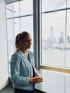 woman with hair pulled back looking out a window at New York City skyline, speaking with a person who is not pictured