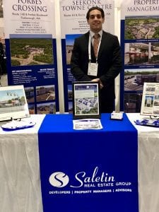 male in a black suit standing at a booth with a blue sign reading "Saletin Real Estate Group"