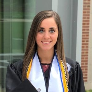 woman with brown hair and blue eyes, wearing black robe with a white collar and various color cords