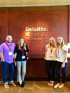 man and three women facing the camera in front of a brown wall with "Deloitte University" written on it