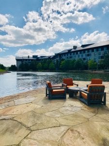 Four brown and orange chairs around a table in front of a blue lake. The lake is in front of a tall building with windows