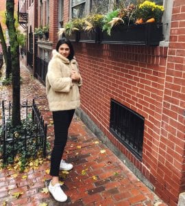 Woman in black pants and brown coat looking at the camera while standing on a sidewalk in front of a brick wall