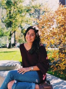 girl with black hair looking at the camera wearing a red sweater and blue jeans. sitting down on a grey bench on a sunny day in front of a green lawn