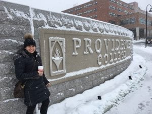woman in a black coat and hat holding a coffee standing in front of the Providence College sign in Huxley Circle in the snow