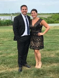 man in a black suit standing with his wife who is wearing a black dress; outside on green grass