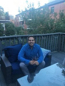 man sitting on a blue chair holding a glass of white wine; outside on a rooftop