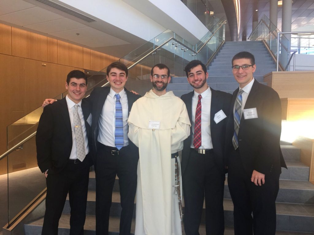4 students in suits posed with a dominician friar in the Ryan Center.