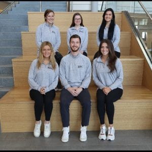 Fellowship students seated on wooden steps inside the Ryan Center for Business Studies. Top row, from left: Chloe Hohmann '23, Madison Palmieri '22, Chae McConaghy '22 Bottom row from left: Sara Dever '22, Ethan Foley '22, Elie Delaney '22