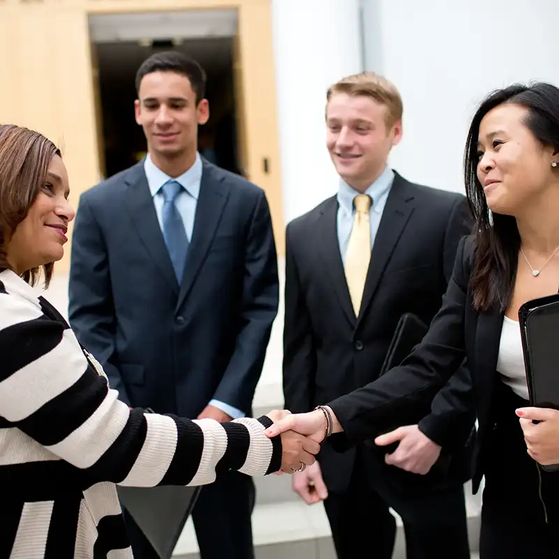 A female student dressed in a business suit and carrying a portfolio shakes hands with a career recruiter while two young men in suits look on.