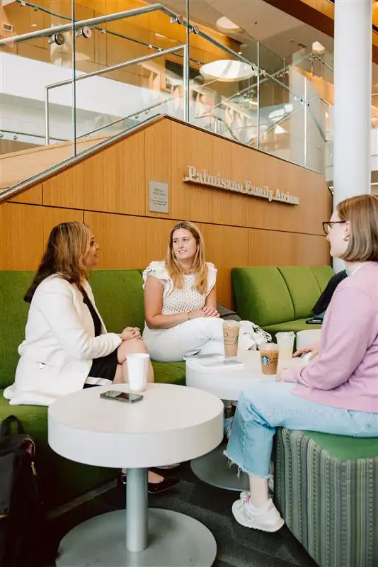 Professor Arati Kale sits with two students in the Palmisano Family Atrium, engaged in a casual discussion around white tables with coffee cups