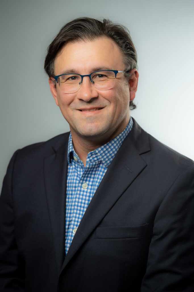 A close up photo of Professor Christopher Lyddy who teaches Management in the Providence College School of Business. He is wearing a dark colored suit with a blue checkered shirt.