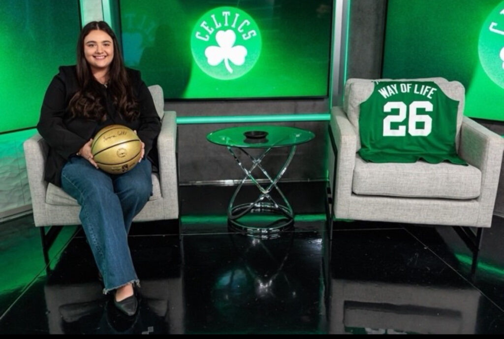 Jenna Cobb '24 at her current job at the Boston Celtics. She is holding a golden basketball and sitting in a gray chair with a Celtics jersey displayed on her right and the Celtics logo displayed in the back.