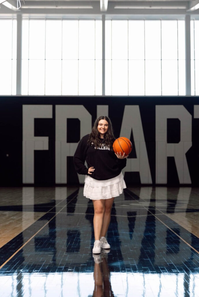 Jenna Cobb '24 standing in the one of the basketball courts on the Providence College campus. She is holding a orange basketball and she is wearing a black sweater with a white skirt. "Friar" appears behind her.