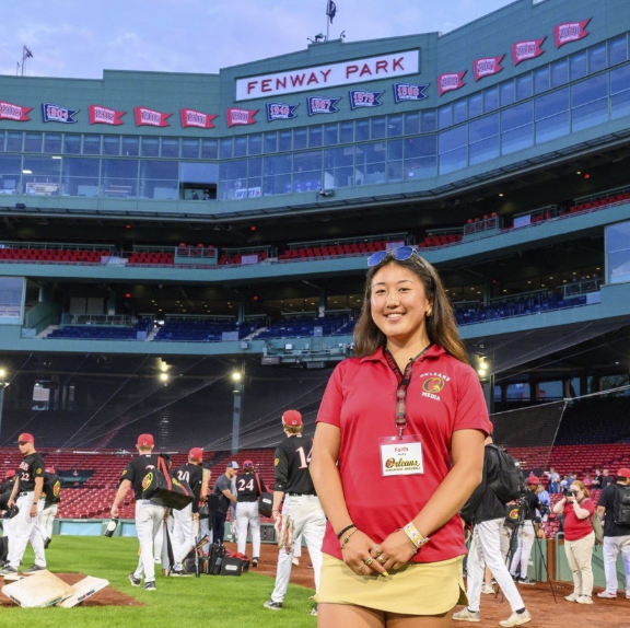 Faith Clark at her summer internship. She is wearing a red shirt, and a tan skirt. She is on the Boston Red Sox field and behind her is a Fenway Park sign.
