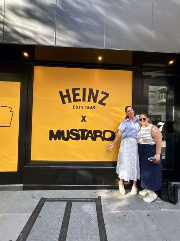 Maddie Frick and a friend standing in front of a Heinz x. Mustard yellow sign.