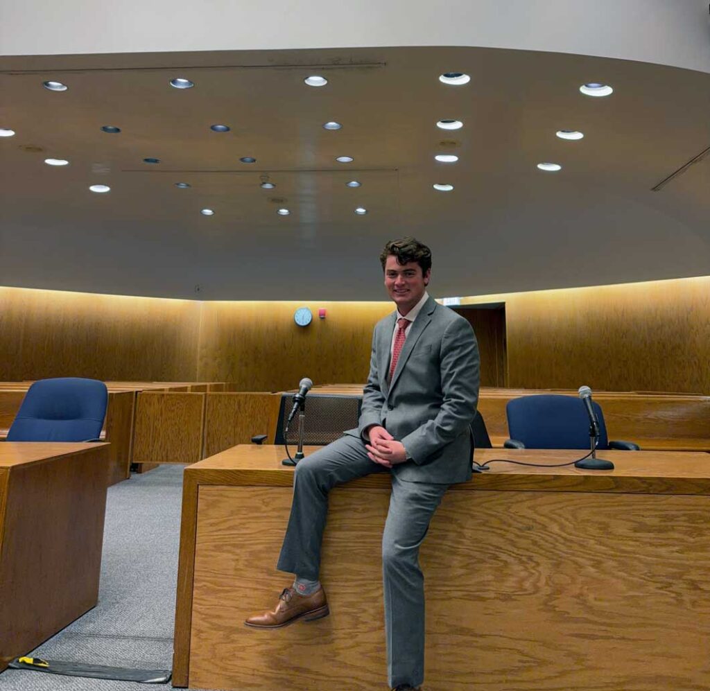 Mikey Stamatis wearing a gray suit as he sits on a desk at the New Haven Superior Court.