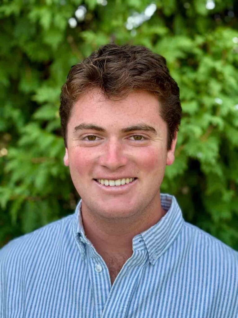 Mikey Stamatis wearing a blue and white striped button down in front of green bushes.