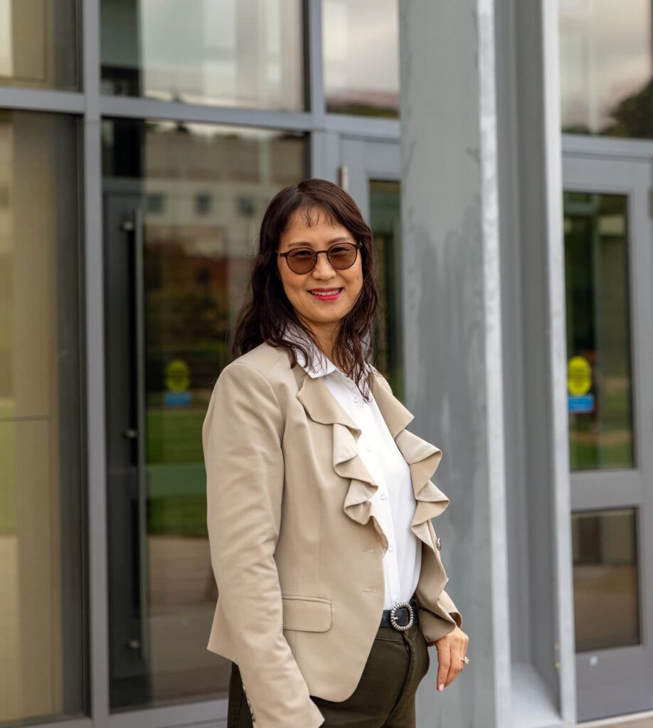 Professor Joy Wilkinson standing outside of the glass doors of the Ryan Center for Business Studies. She is wearing sunglasses and a tan ruffled jacket.